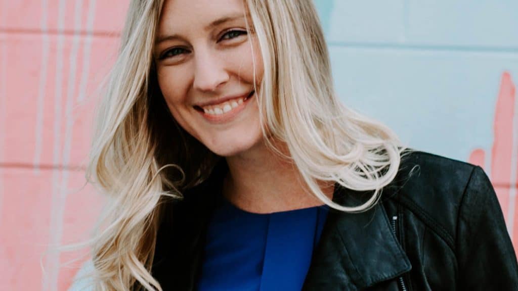 Young, blonde-haired woman smiling and standing in front of a pink and blue mural