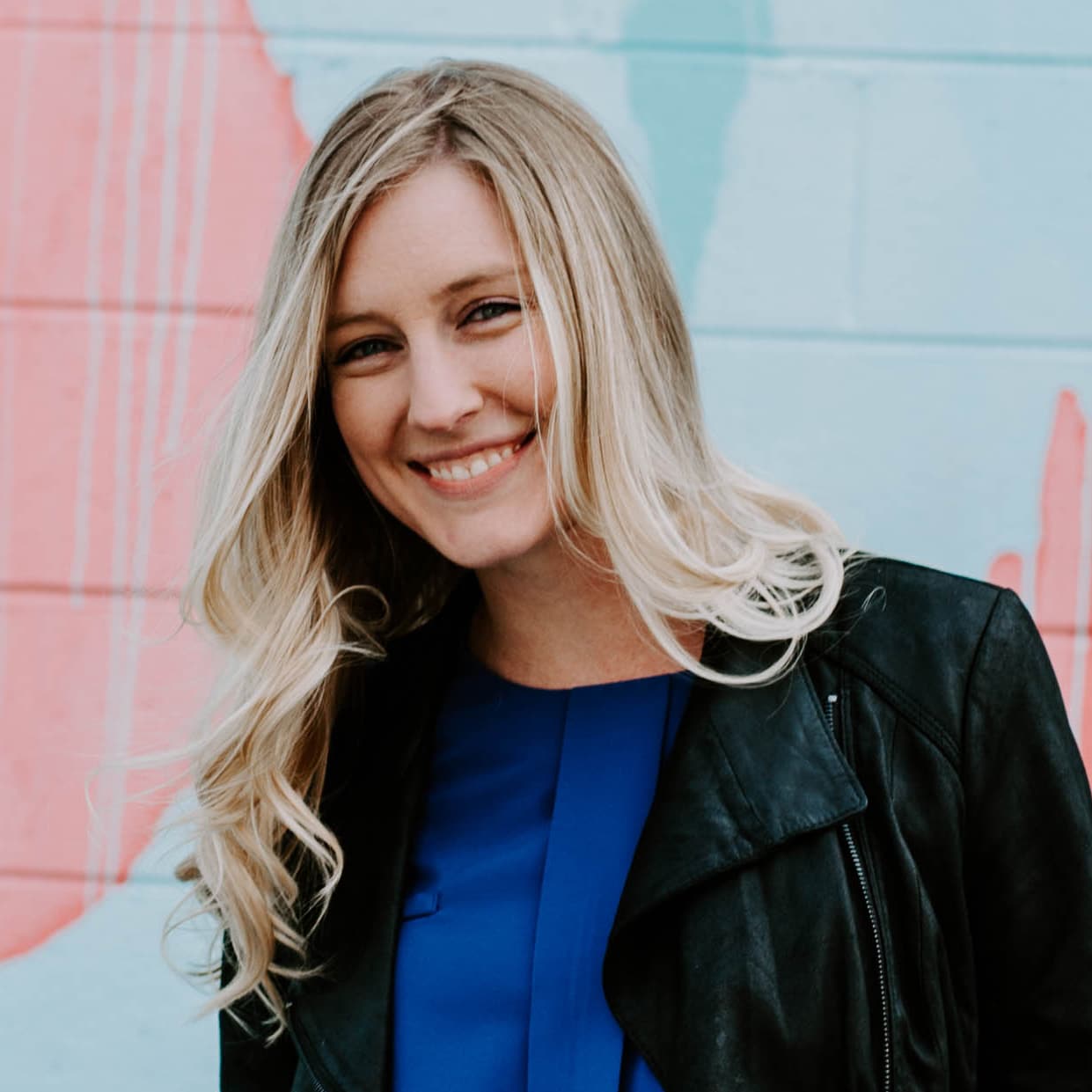 Young, blonde-haired woman smiling and standing in front of a pink and blue mural