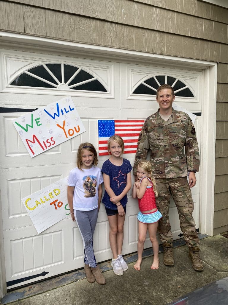 A dad in military uniform with three young girls standing before homemade signs