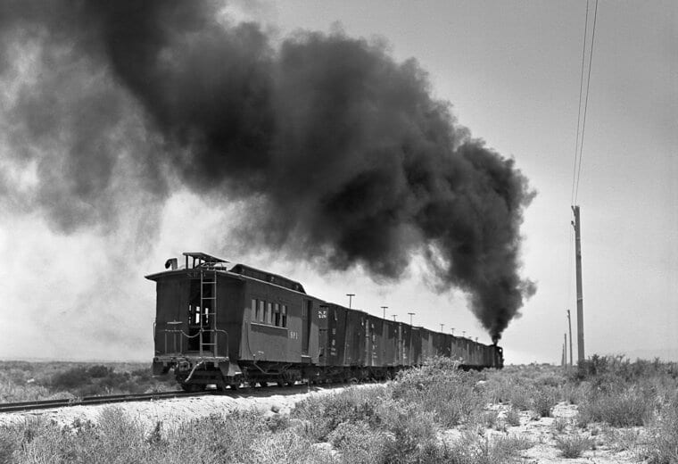 Black and white image of a old train traveling through flat landscape with a pillar of black smoke