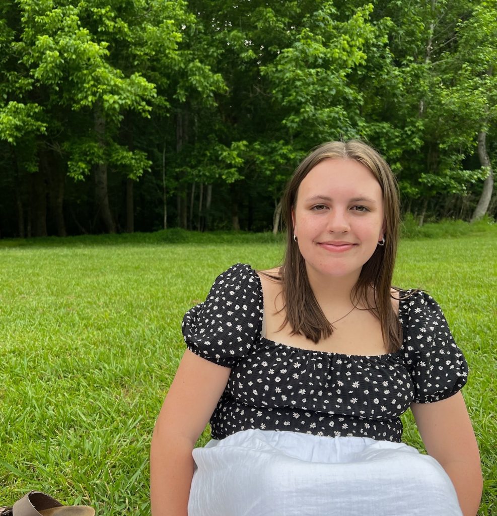 Young woman with straight brown hair sitting in the grass with a white blanket over her legs. Lush green trees line the background.