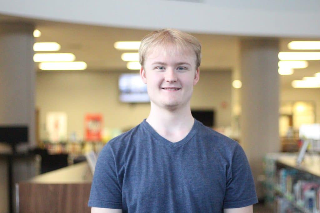 Blonde haired young man in a blue shirt, smiling and looking at the camera.