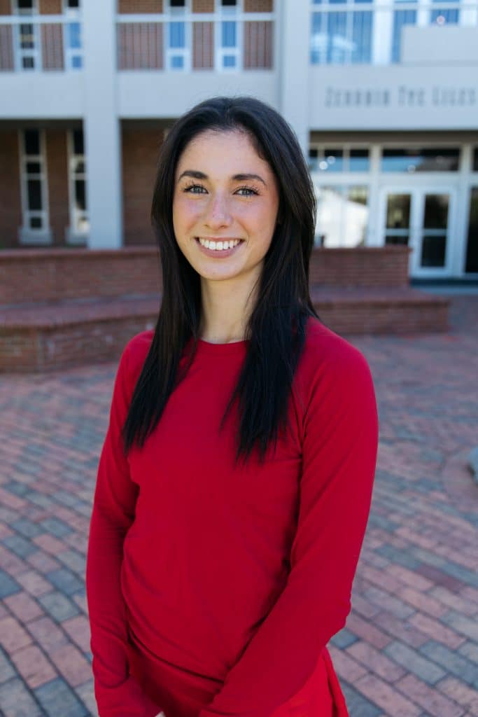 Young Hispanic high school student. She's got long straight dark hair, is wearing a red sweater, and is standing outside her school, smiling at the camera.