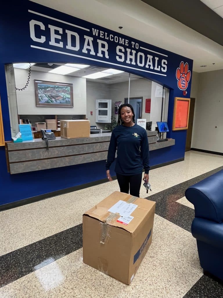 Young Black woman, high-school aged, wearing a blue athletic top and pants, standing behind a large box that she is delivering to Cedar Shoals school.