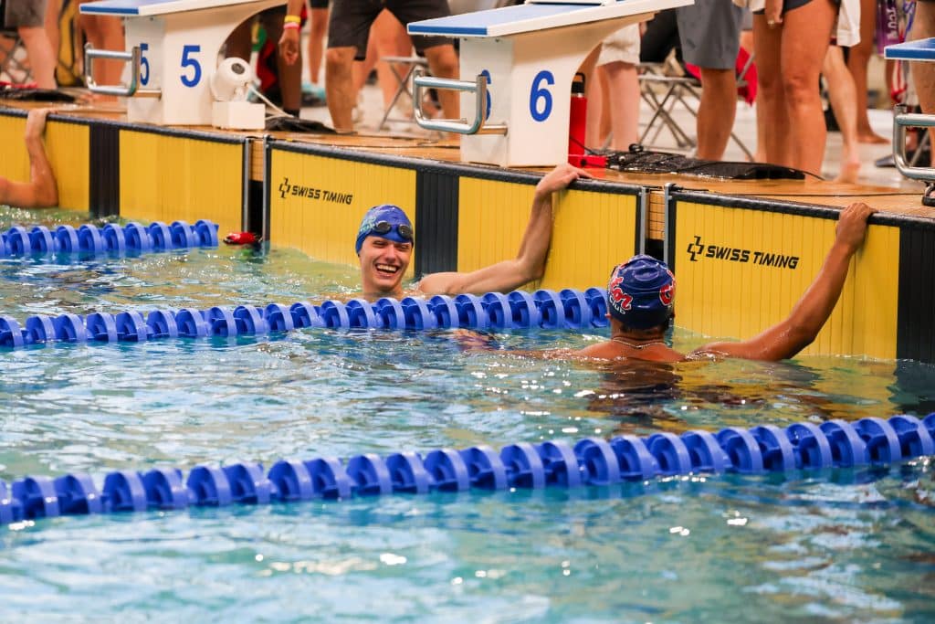 Young man in a pool, wearing a swim cap and smiling at another swimmer after finishing a swim competition.