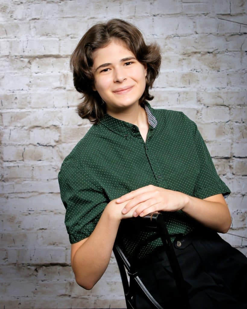 Young adult with brown hair, wearing a green, collared shirt. They're sitting sideways in a chair, leaning against the back of it.