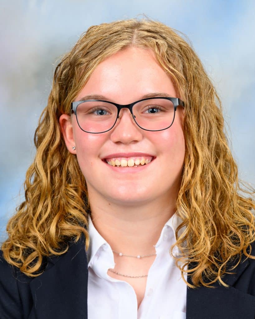 School photo of a young white female student with blond curly hair and glasses. She's wearing a white button up shirt and black blazer and smiling.