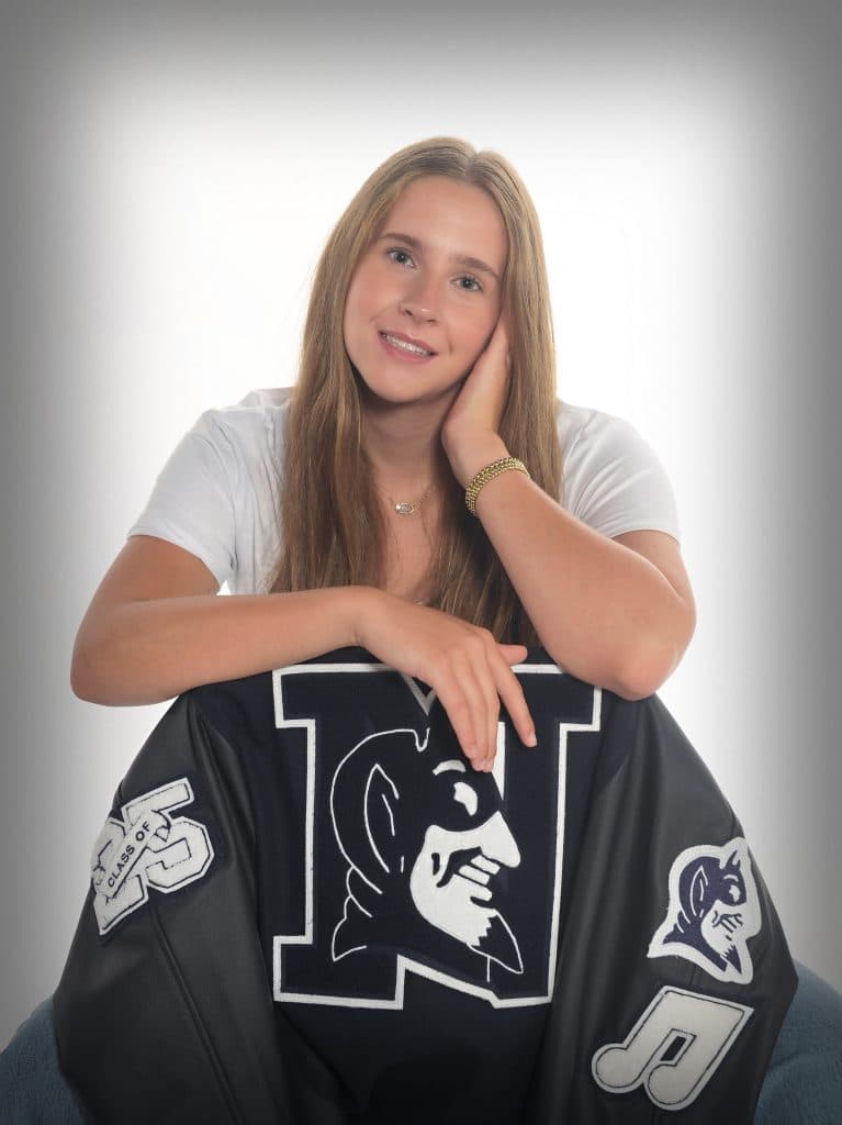 White female high school student with long, straight blonde hair, sitting backwards in a chair draped with her school jacket. She's resting her arms on the chair's back and her head is tilted to one side.