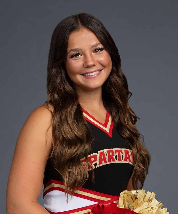 Young white high school female with long brown hair, wearing a cheerleading outfit. She's looking at the camera with head slightly tilted to her right while smiling.