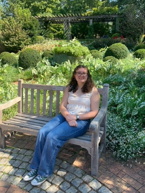 Young white high school student sitting on a bench in a garden. She's got shoulder-length brown hair and glasses and is wearing a light-colored top and blue jeans.