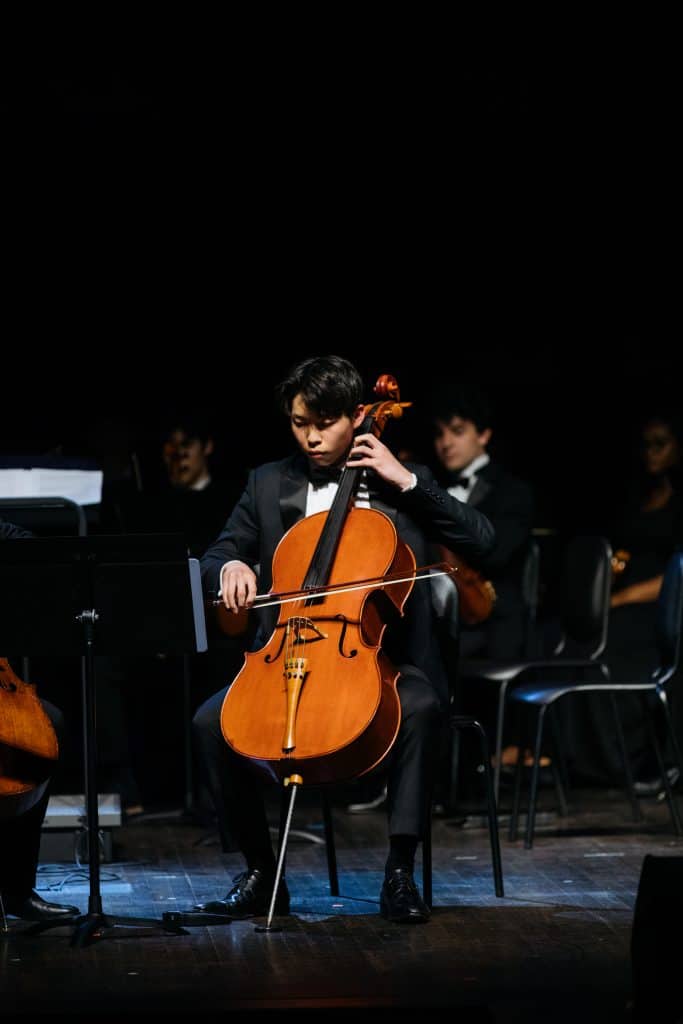 Young Asian male wearing a suit, on stage with an orchestra as he plays the cello.