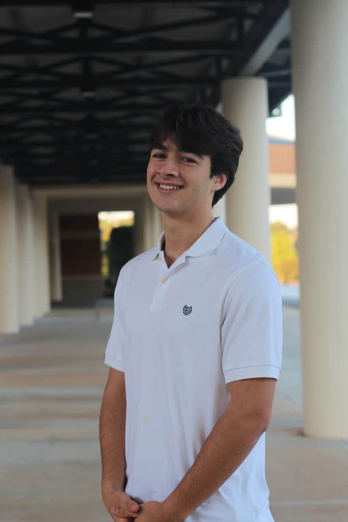 Young Hispanic male high school student wearing a white short-sleeved shirt. He's standing outside at his school, turned at an angle towards the camera and smiling.