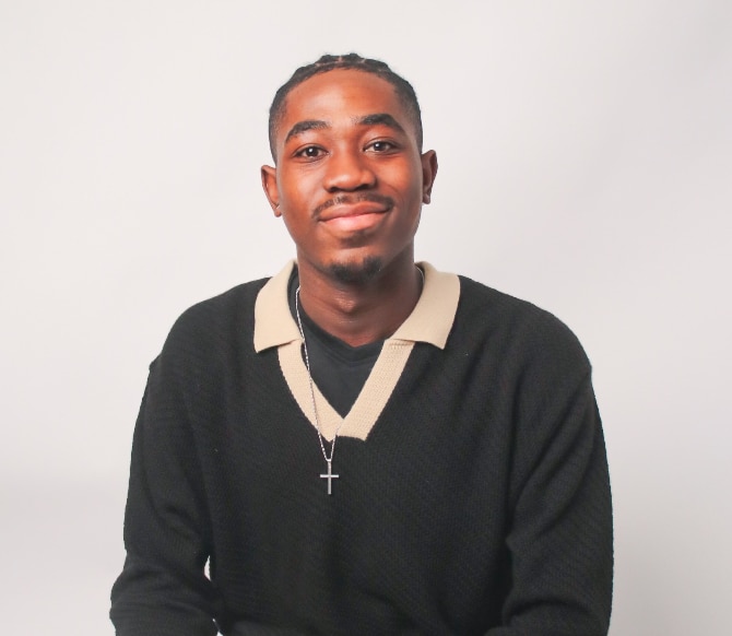 Young Black man wearing a dark pullover shirt with white color and a cross necklace, sitting against a white background. He's looking directly at the camera and smiling.