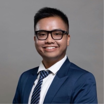 Young Asian male wearing white shirt and blue suit and tie. He's standing against a light-colored background, wearing glasses and smiling.