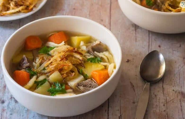 Bowl of soup with chicken and vegetables on a wooden table next to a spoon