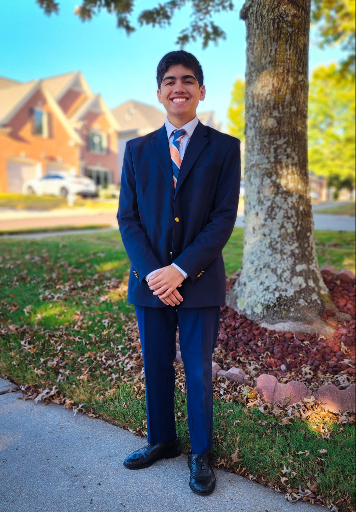 Asian male high school student, wearing a suit. He's standing outside with trees and houses behind him, and he's smiling at the camera.