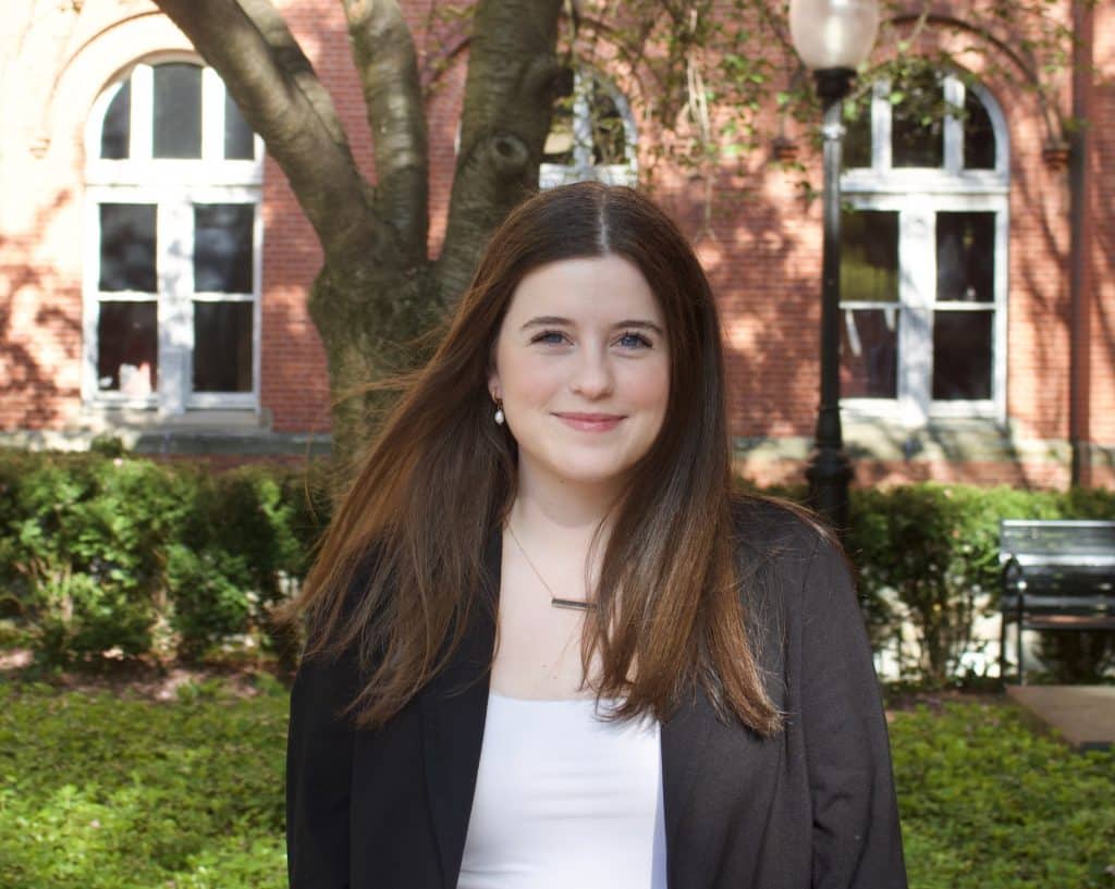 Female college student with long brown hair, wearing a white top and dark-colored jacket. She's standing in front of a tree with a school building behind it, and she's smiling.