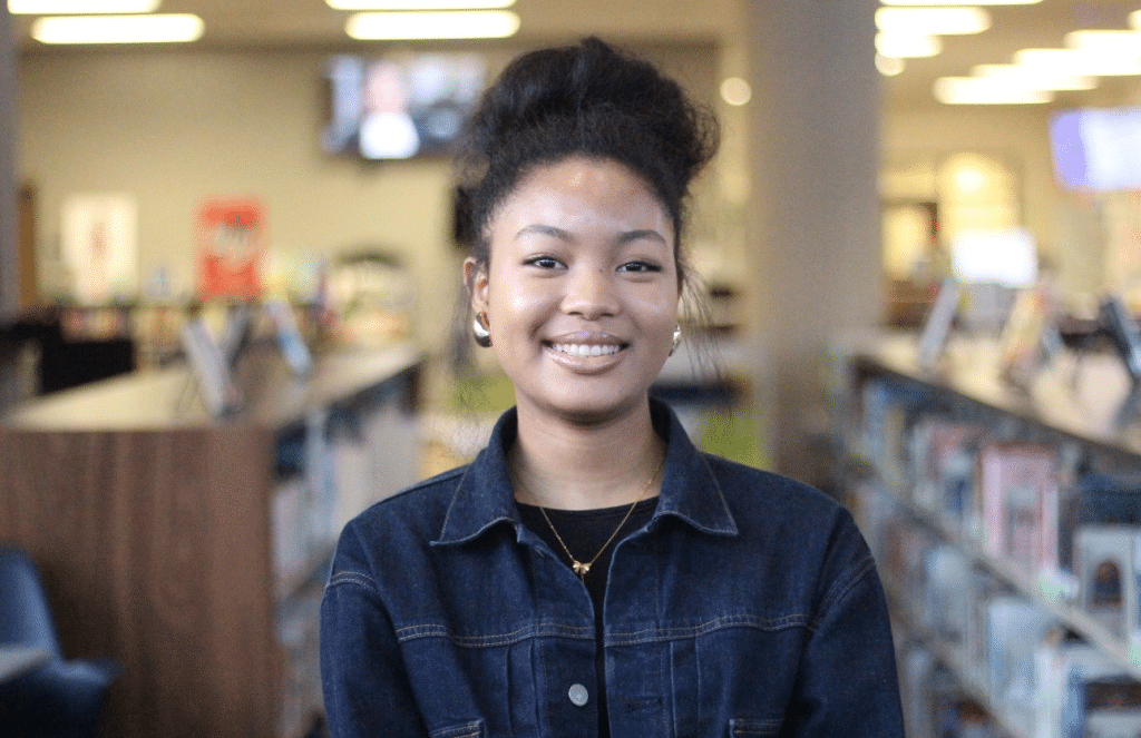 Young Black-Asian woman wearing a dark blue top. She's standing in a library, her hair is pulled up and she's smiling at the camera.