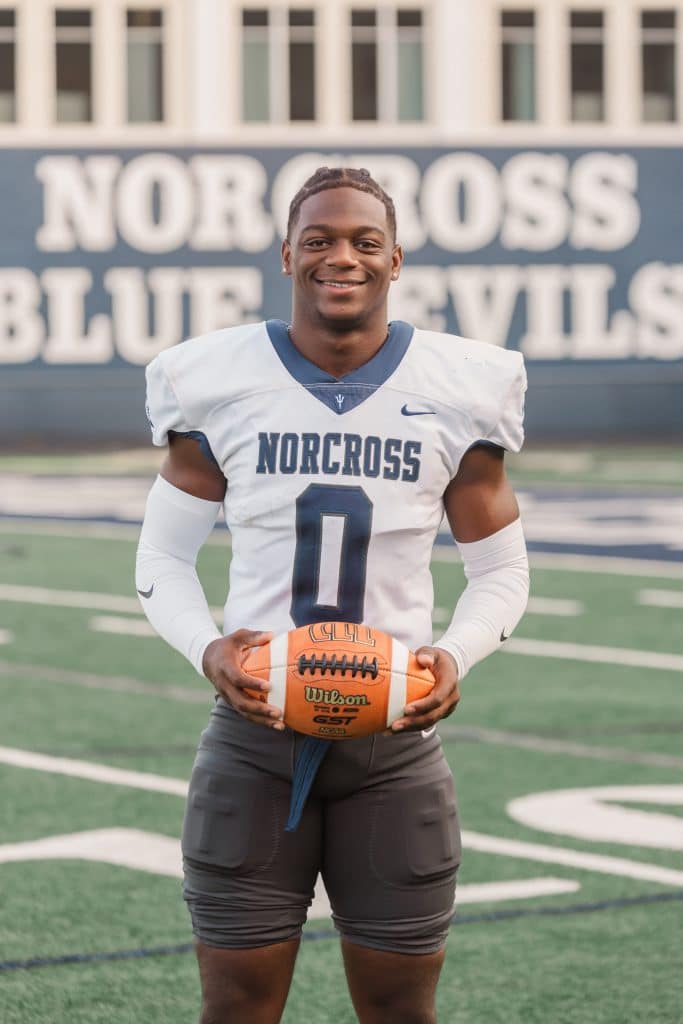 Black male high school football player, in practice uniform, standing on the football field holding a football and smiling at the camera.