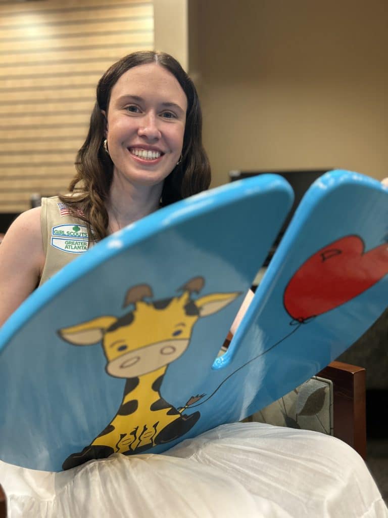 Young white woman with shoulder-length brown hair. She's seated and wearing a white skirt and Girl Scout vest while smiling and holding a "lily pad seat" painted blue with a yellow and brown giraffe on it.