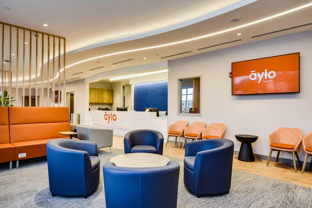 Healthcare center waiting room with modern, clean design, blue chairs, orange sofa, a reception desk and a tv screen on the wall showing the Aylo Health logo