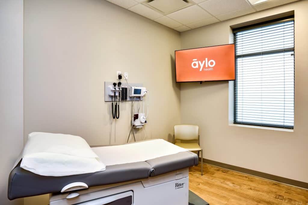 Exam room in a new healthcare facility, with exam table, chair, window and tv screen showing the Aylo Health logo