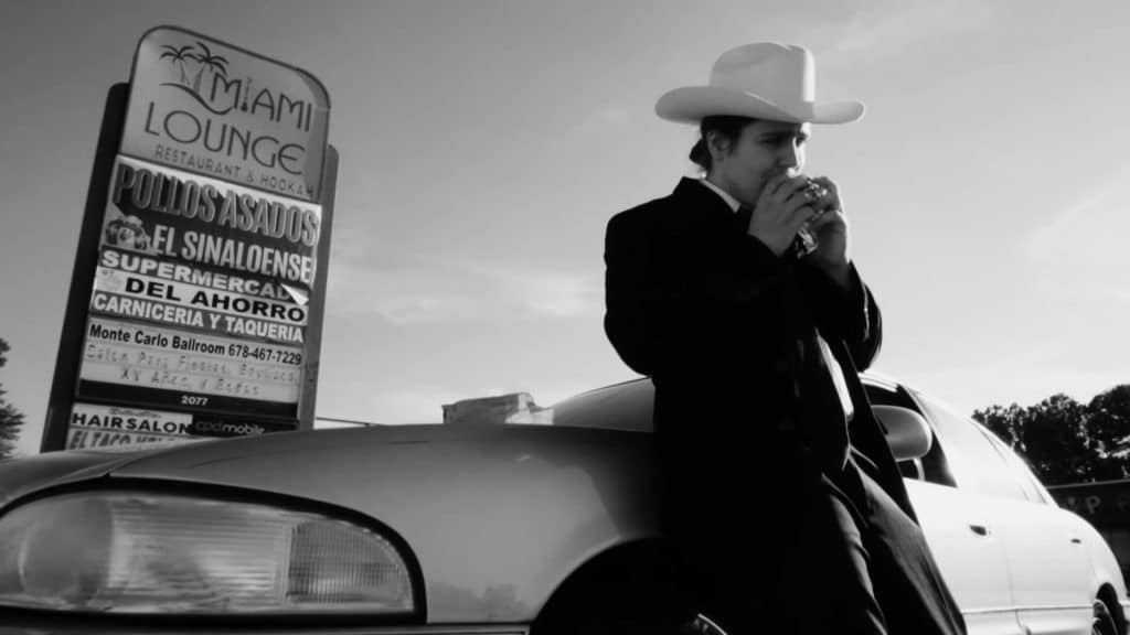 Black and white shot of a man wearing a suit and a white cowboy hat leaning against a car in the parking lot of a restaurant and retail center.