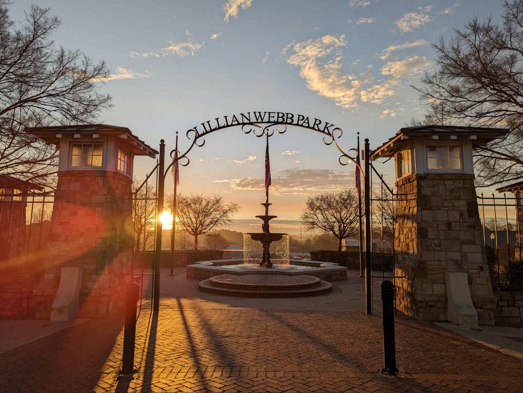 Sunrise shining through a large arched sign for Lillian Webb Park. The sky is light blue clouds and there is also a fountain and other structures in the mid-ground.