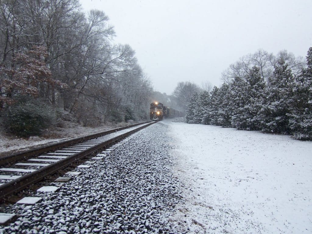 Train in the distance on a railroad track lined with tall green trees. There's a grey, overcast sky above and light snow on the ground.