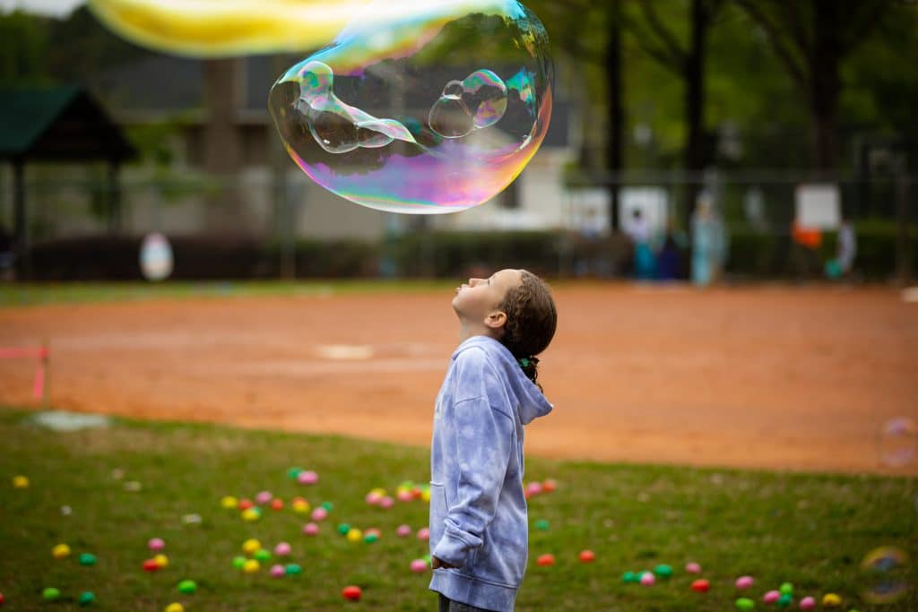 Young child with a light blue/purple shirt standing in a park field looking up at large bubbles floating above them.