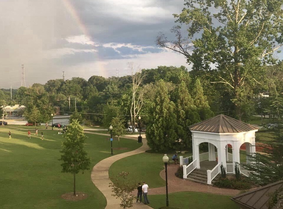 Blue sky with clouds and rainbow over a green space park with trees and white gazebo.