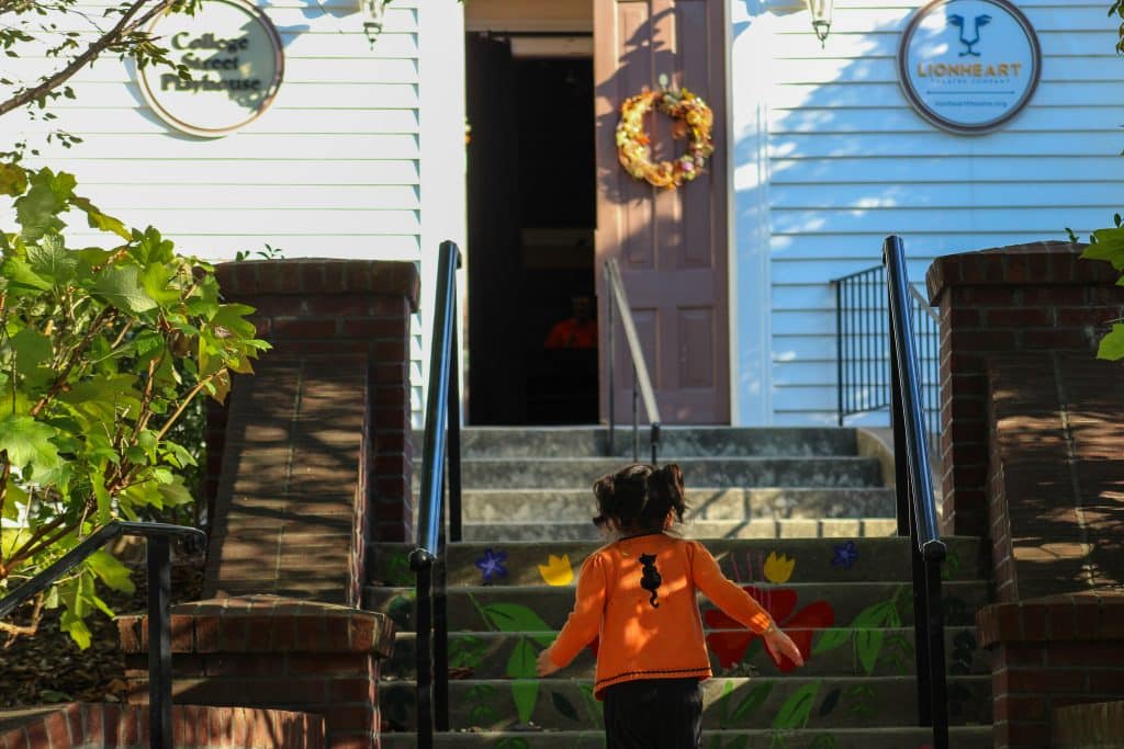 Small child in orange shirt running up the steps to a white wooden theater building