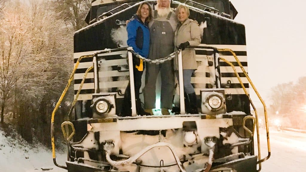 A photo of the front of a train, stopped on snowy tracks. Two women and a train engineer that looks a lot like Santa Clause stand at the front posing for the camera.