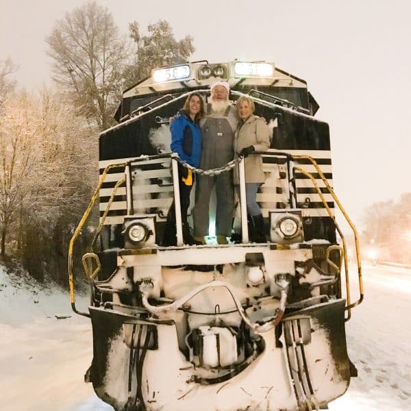 A photo of the front of a train, stopped on snowy tracks. Two women and a train engineer that looks a lot like Santa Clause stand at the front posing for the camera.