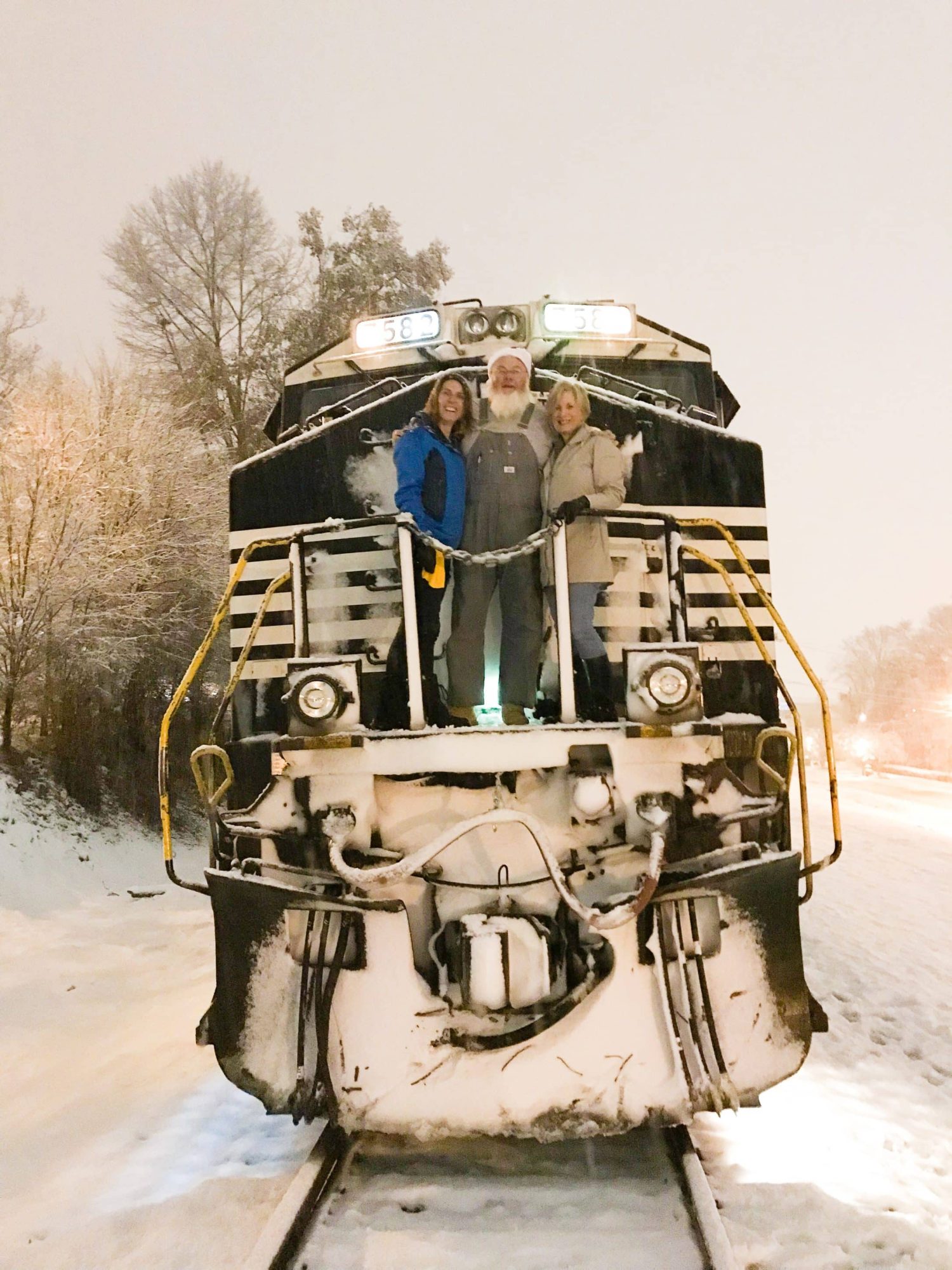 A photo of the front of a train, stopped on snowy tracks. Two women and a train engineer that looks a lot like Santa Clause stand at the front posing for the camera.