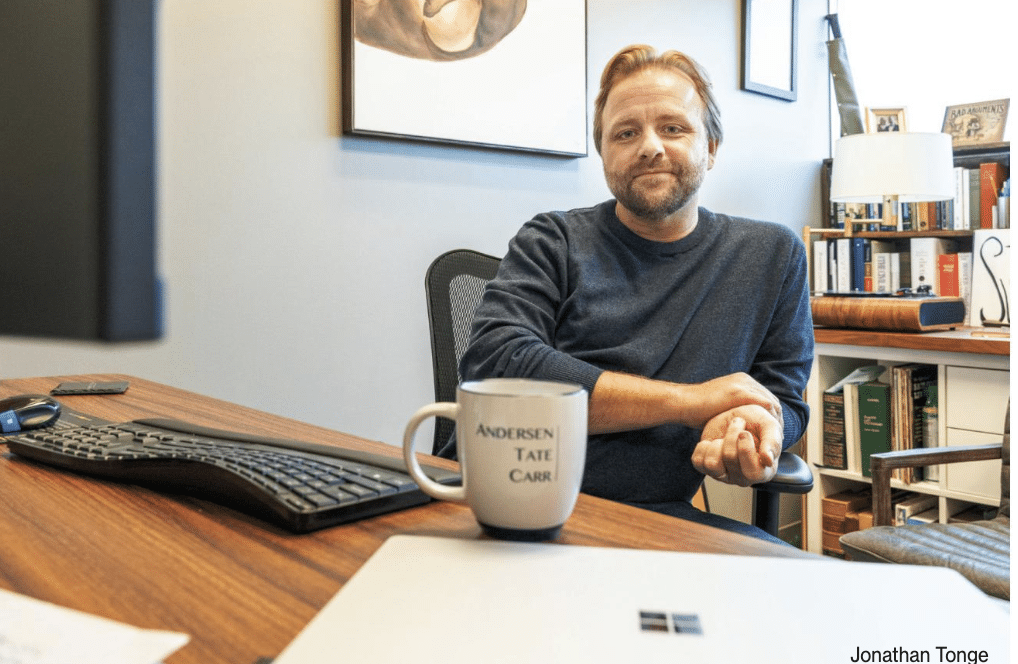 Man in casual clothing sitting at a desk with computer keyboard and mug of coffee in front of him.