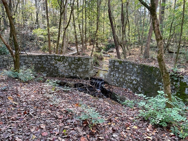 A wooded area with stone-sided boardwalk over a small stream in fall