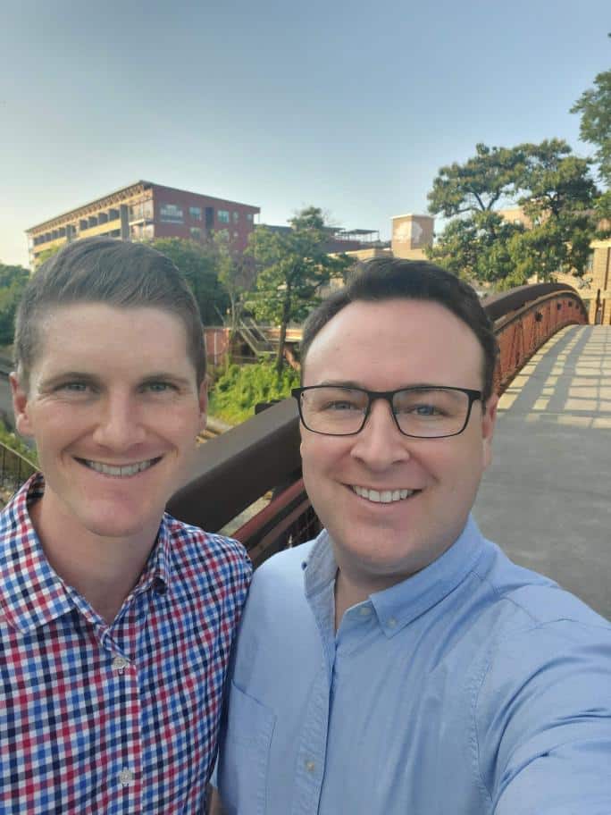 Two white men, smiling and taking a selfie outdoors. The man on the left is wearing a small-checked button up shirt. The one on the right is wearing a light blue button up shirt and glasses.