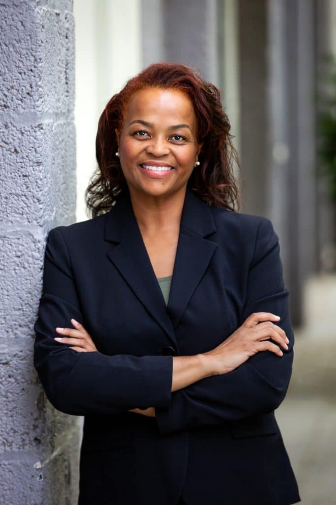 A Black woman with shoulder-length hair, wearing a dark suit. She's leaning against a grey wall with arms crossed in front of her, smiling.