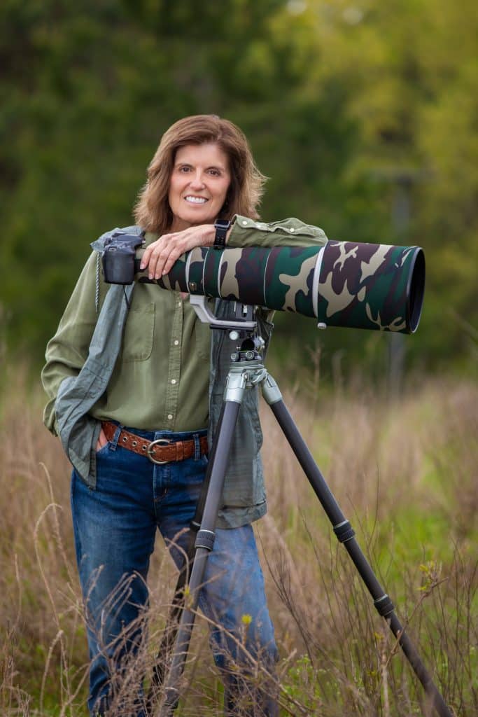 A woman with shoulder-length brown hair, wearing green top and light jacket and jeans, standing in an open field and leaning against a large telephoto camera on a tripod. There's a line of trees in the background behind her.