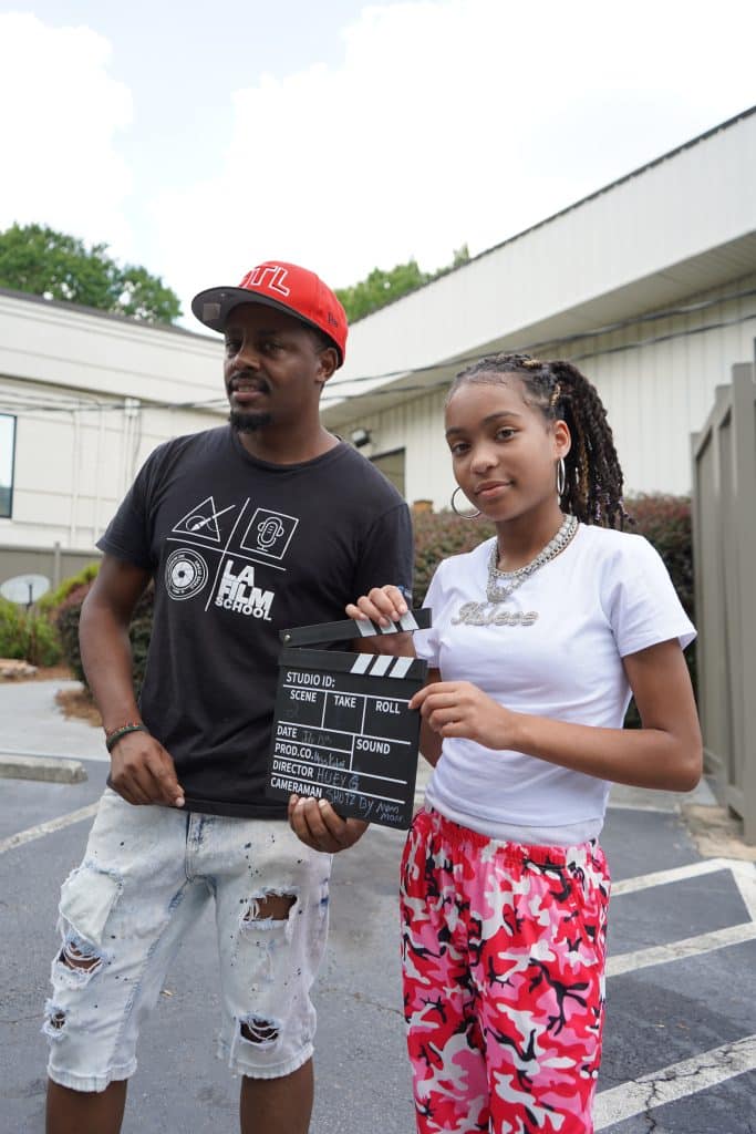 A Black man wearing a red cap, black tshirt and shorts standing next to a Black, teenage girl wearing a white tshirt and red print pants as they work on a video shoot.