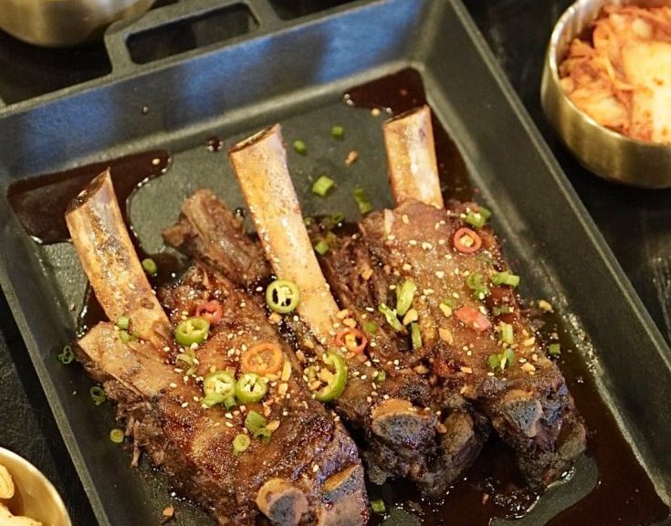 Korean BBQ ribs and side dishes served in separate plates on a black table. The ribs are covered with tiny sliced red peppers and green onions.