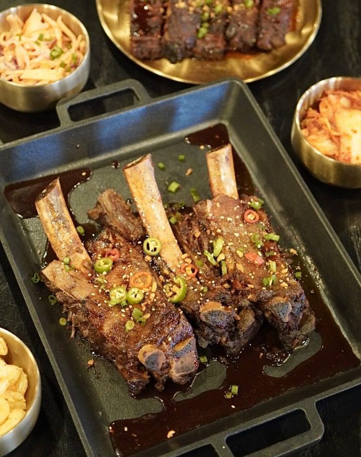 Korean BBQ ribs and side dishes served in separate plates on a black table. The ribs are covered with tiny sliced red peppers and green onions.