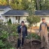 Two people with shovels standing in the front yard of a white split-level home next to a tree they've just planted.