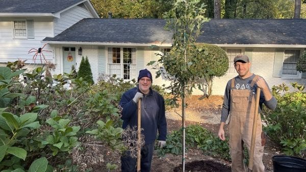 Two people with shovels standing in the front yard of a white split-level home next to a tree they've just planted.