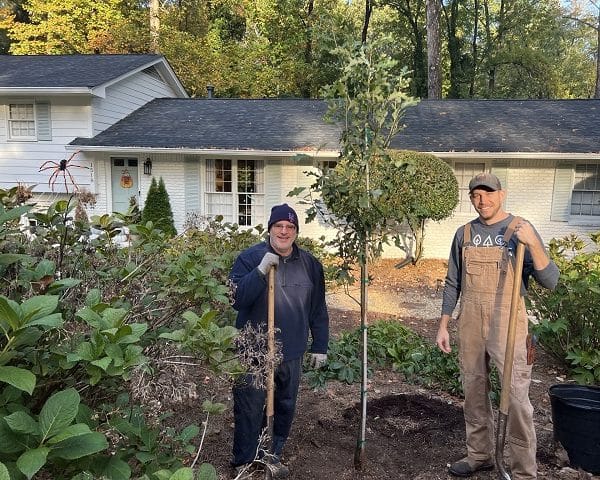 Two people with shovels standing in the front yard of a white split-level home next to a tree they've just planted.