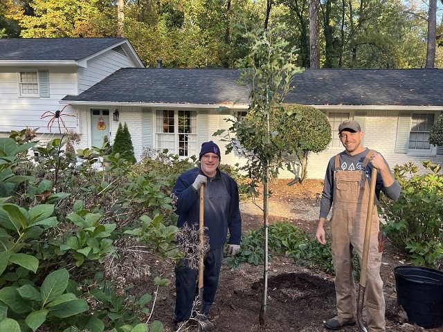 Two people with shovels standing in the front yard of a white split-level home next to a tree they've just planted.