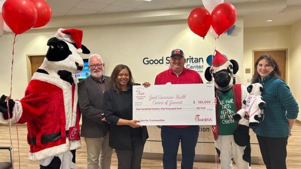 A group of people from Good Samaritan Gwinnett and Chick-fil-A standing in the waiting area of GSG, holding a giant check. There are red and white balloons, a Chick-fil-A cow mascot in a Santa suit. The check represents a $125,000 donation grant to GSG.