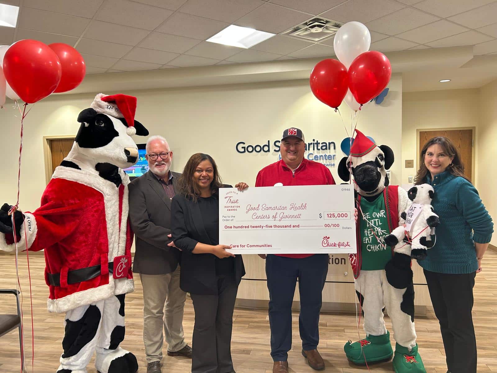 A group of people from Good Samaritan Gwinnett and Chick-fil-A standing in the waiting area of GSG, holding a giant check. There are red and white balloons, a Chick-fil-A cow mascot in a Santa suit. The check represents a $125,000 donation grant to GSG.