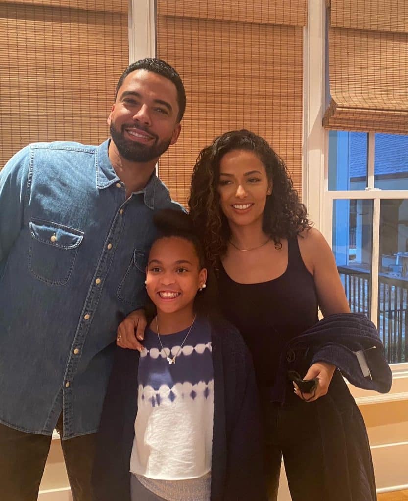 A Black man and woman standing with a young, Black female. Their arms are around each other and they're smiling at the camera on the set of the Lifetime movie, Stalked by Her Past.